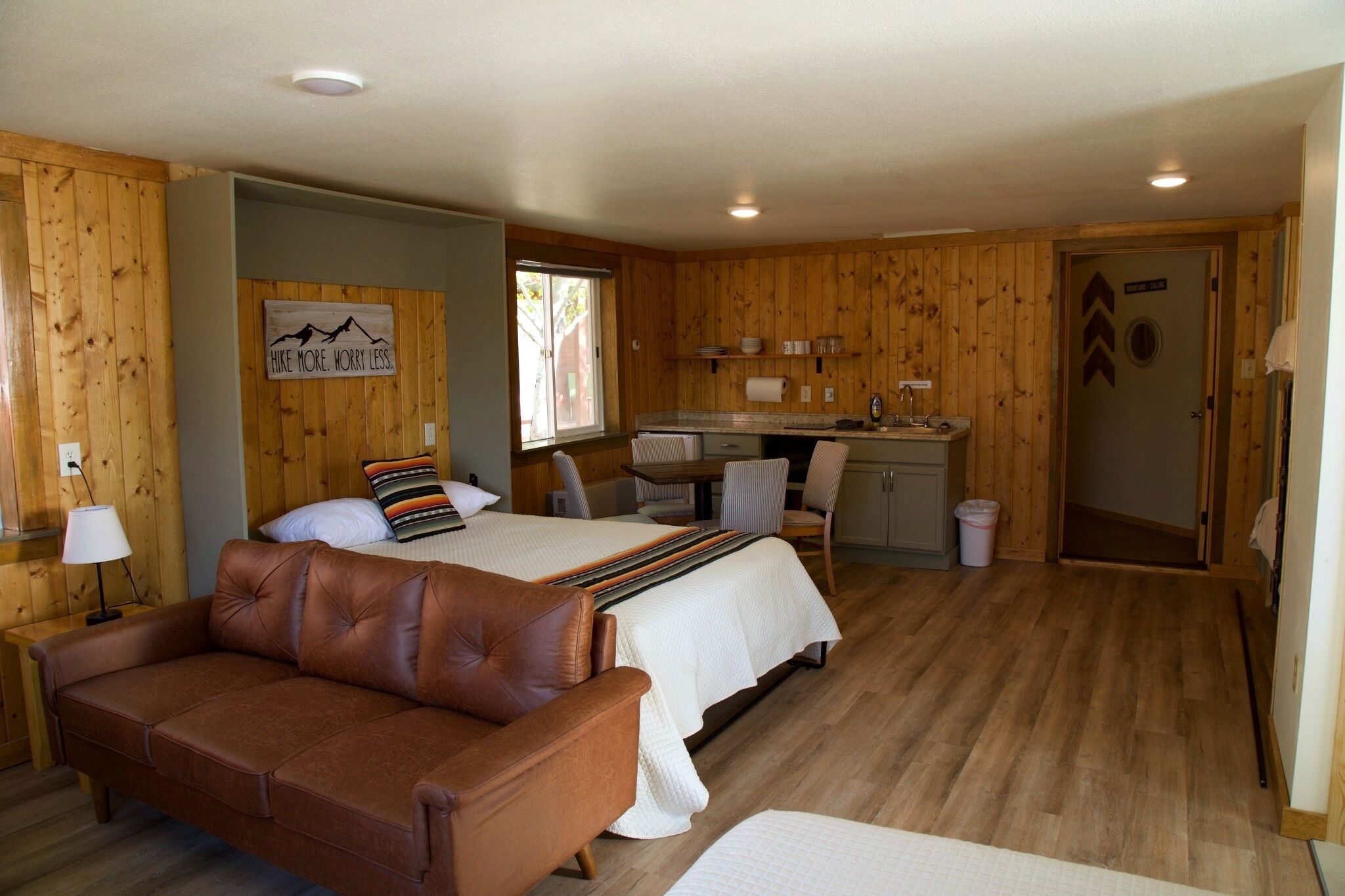 A room with a bed, a brown leather sofa, a small kitchenette, and a wooden wall.