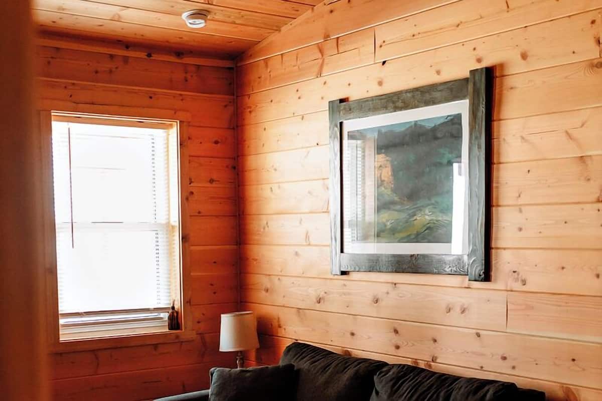 A cozy room with a wooden ceiling, a black leather sofa, a framed picture on the wall, and a window with blinds.