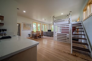 Uma cozinha moderna com bancada branca, piso de madeira, área de jantar com cadeiras e escada com corrimão.