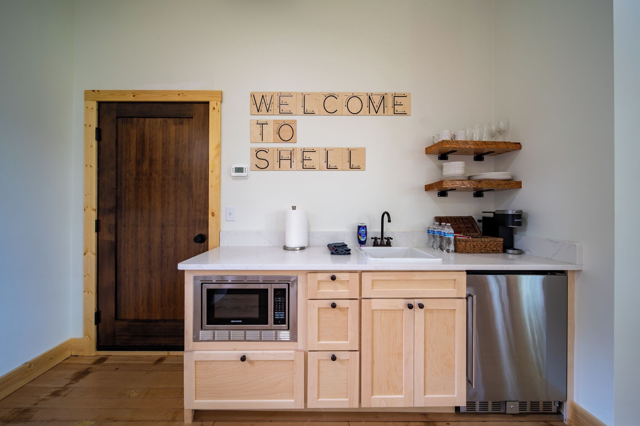 Una cocina con armarios de madera, lavavajillas de acero inoxidable, microondas y un cartel que dice 'WELCOME TO SHELL'.
