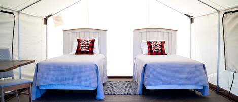 A tented accommodation with two beds, each with a pillow, a small table, and a chair.