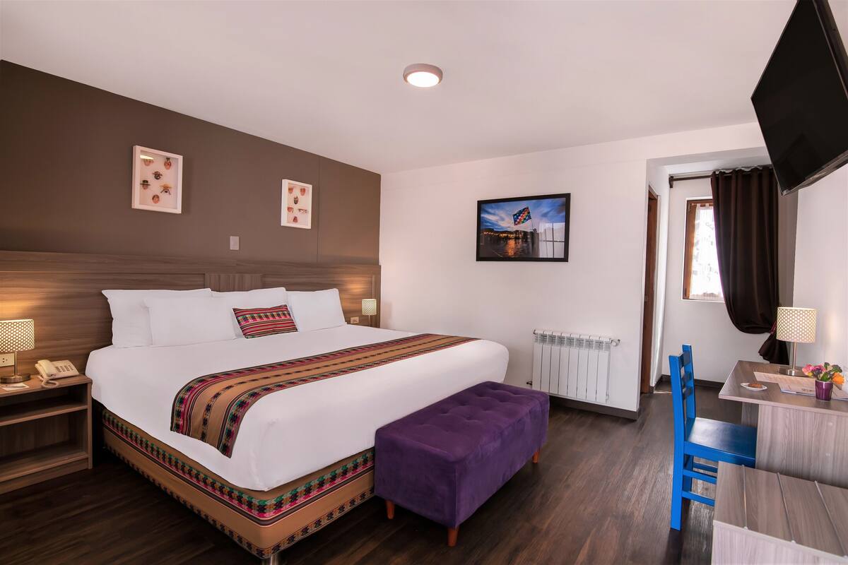Une chambre d’hôtel avec un grand lit, une table de chevet en bois, une petite table avec une lampe, une chaise bleue et une fenêtre avec des rideaux.