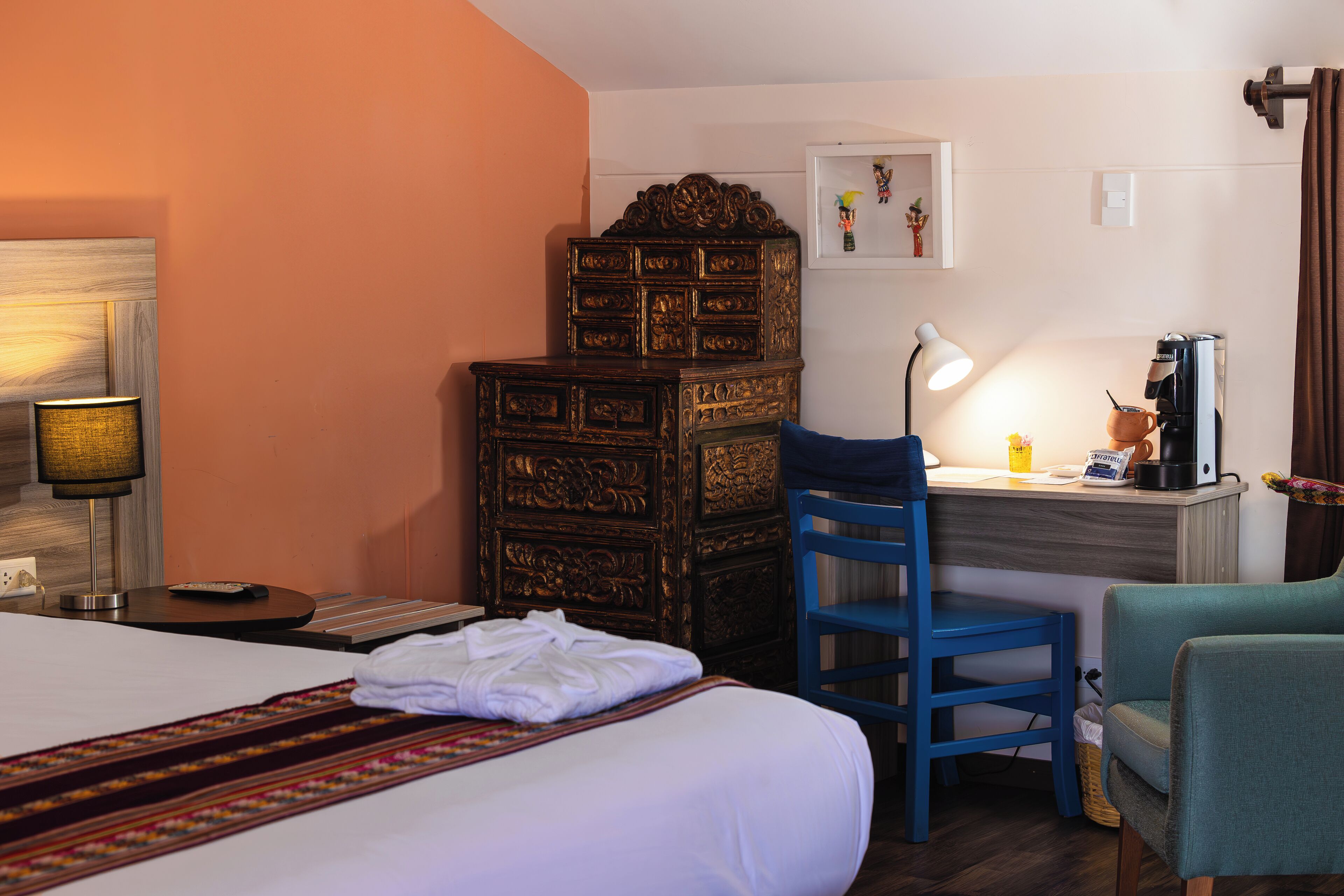 A hotel room with a bed, a desk with a lamp, a coffee machine, and a dresser.