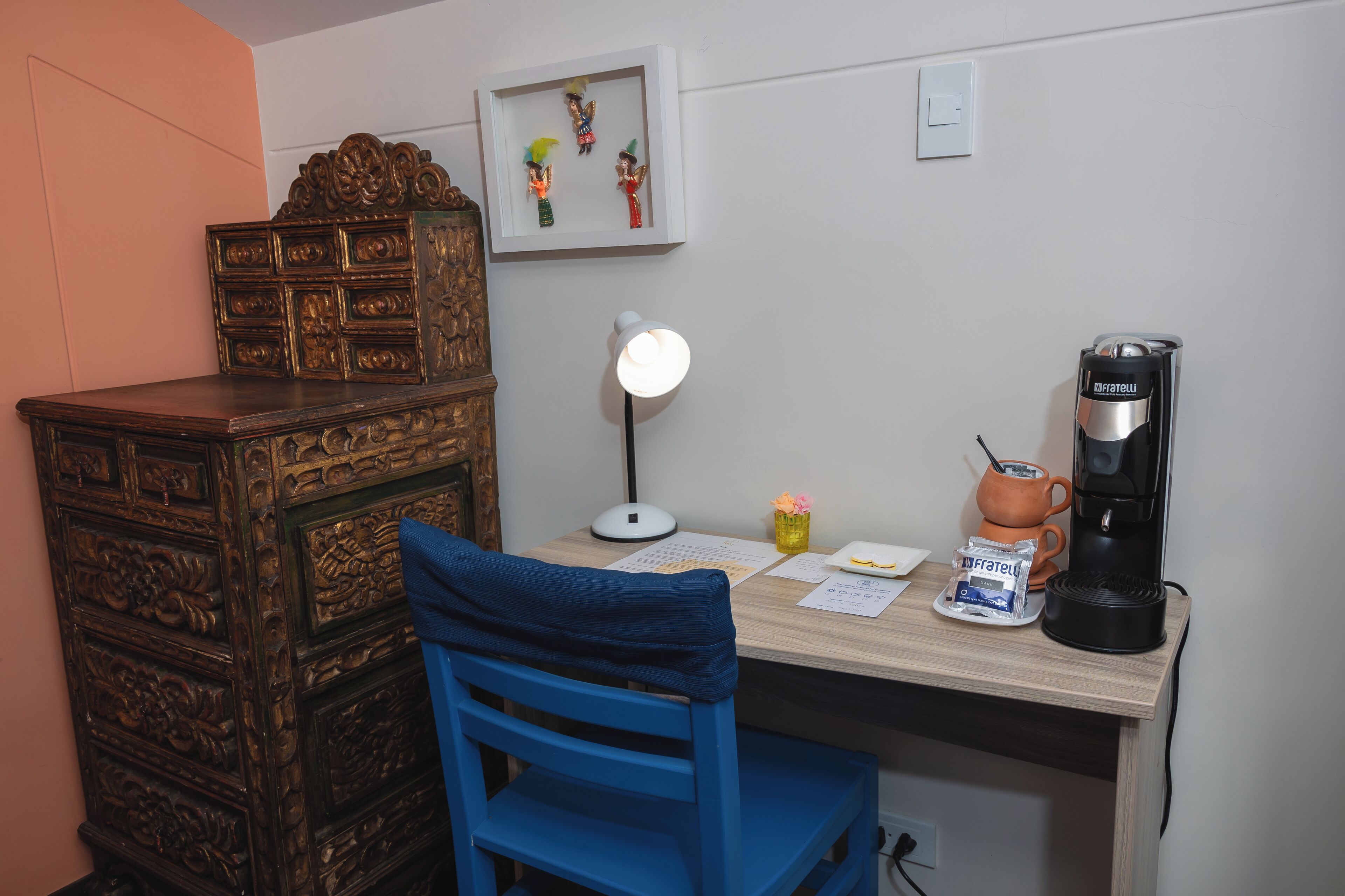 Un bureau en bois, une chaise bleue, une machine à café, une lampe et un tableau encadré accroché au mur.