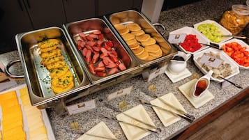 A buffet with various food items including cheese, sausages, pancakes, and fruits.