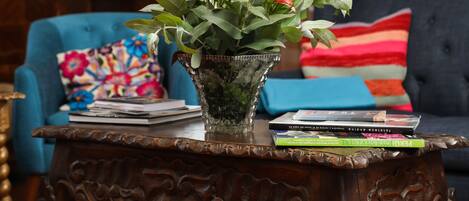 Una mesa de centro de madera con un jarrón de vidrio que contiene flores amarillas, una pila de libros y un cojín colorido sobre un sofá azul al fondo.