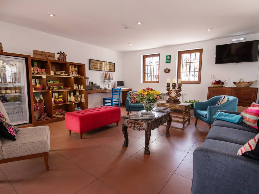 Uma sala de estar ampla com um banquinho vermelho, poltronas azuis, um sofá cinza e uma mesa de centro de madeira. Há uma estante repleta de livros e objetos decorativos, uma televisão fixada na parede e uma janela com vista.