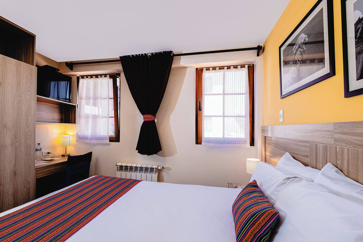 Une chambre d’hôtel avec un lit, un bureau, une chaise, un radiateur et une fenêtre avec des rideaux.