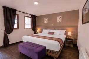 Una habitación de hotel con una cama grande, un banco púrpura, dos mesitas de noche con lámparas y cuadros en la pared.