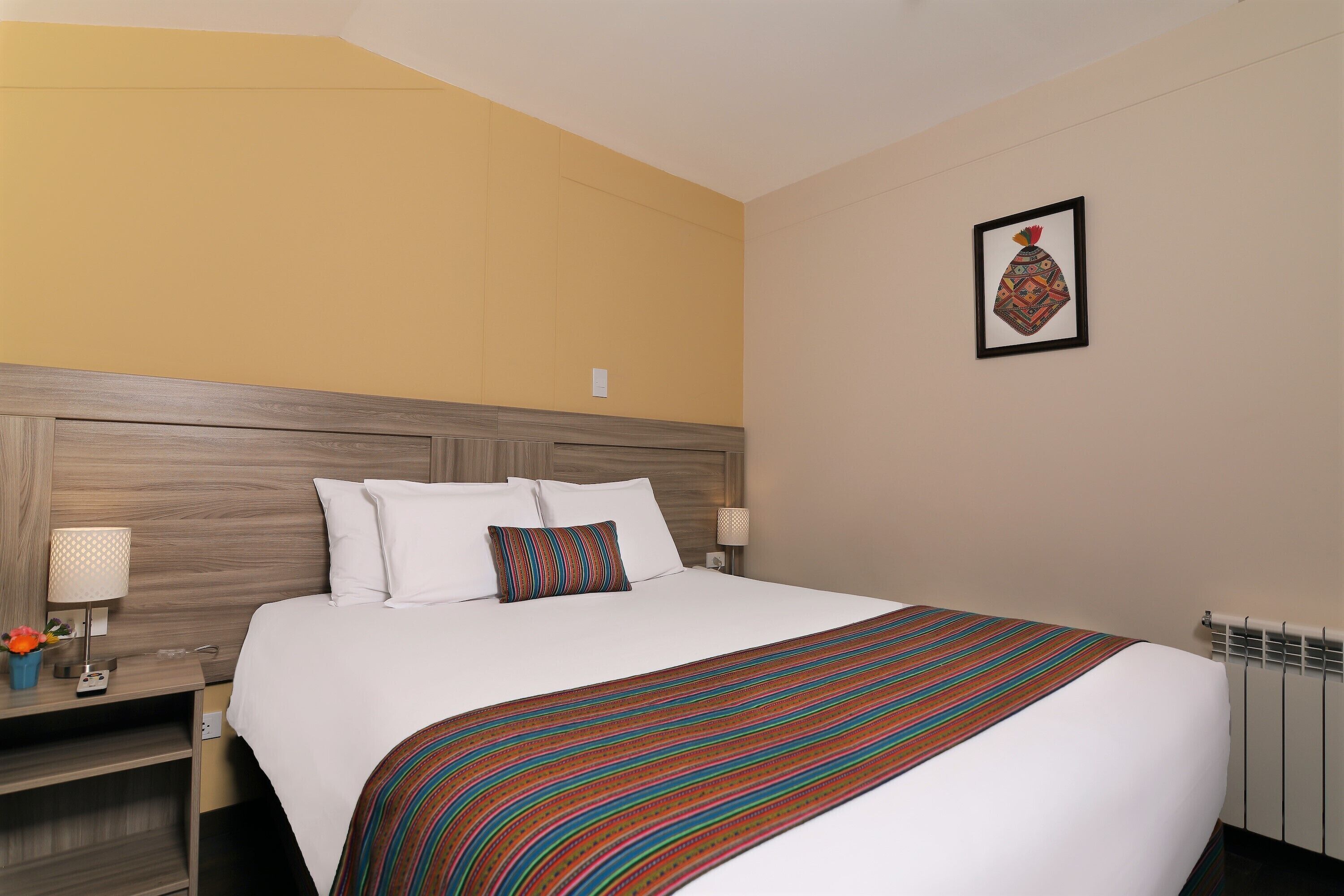 A hotel room with a large bed, a wooden headboard, a colorful striped blanket, white bedding, a small table with a lamp, and a framed picture on the wall.