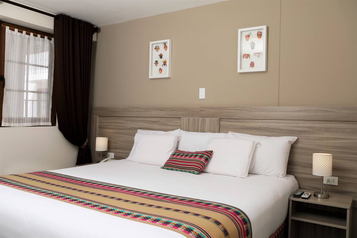 Une chambre d’hôtel avec un grand lit, du linge de lit blanc, un plaid à rayures, deux tableaux encadrés au mur, une table de chevet avec une lampe et une fenêtre avec des rideaux.