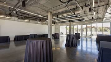 A spacious indoor area with tables covered in tablecloths, a high ceiling with exposed ductwork, and large windows allowing natural light.