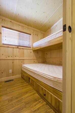 A cabin interior with wooden walls, a bunk bed, a window with blinds, and a vent on the floor. - Sunset Lakes RV Resort (Hillsdale)
