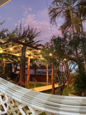Una hamaca bajo una pérgola de madera con luces colgantes, rodeada de follaje y un cielo despejado.