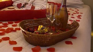 Una cama con una colcha roja y blanca con diseño de corazones, una cesta con pétalos de rosa, una botella de licor y un vaso sobre la cama.