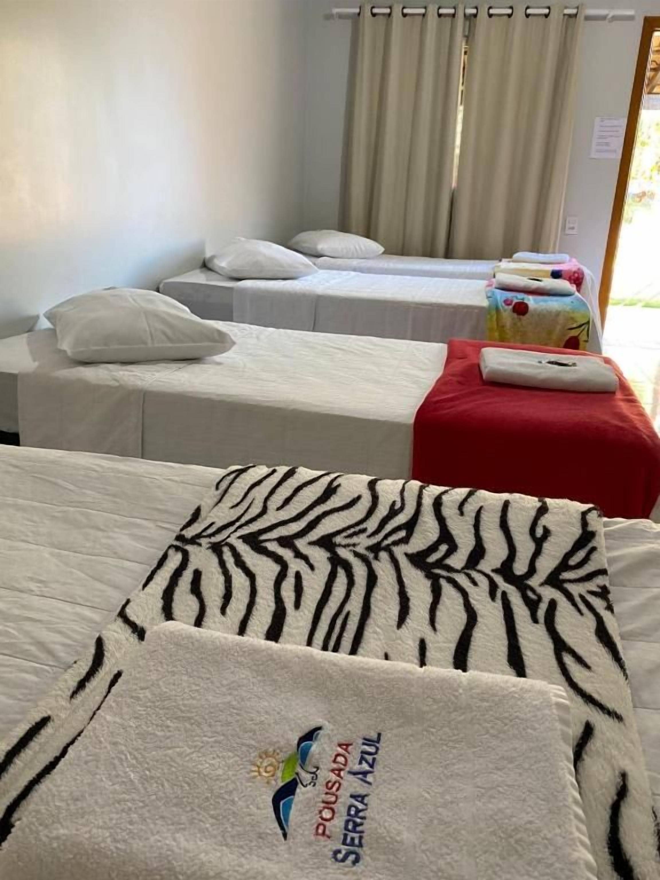 Habitación de hotel con dos camas individuales, cada una con cubrecama blanco y manta roja. Se observa una ventana con cortinas y una pequeña mesa con mantel rojo.