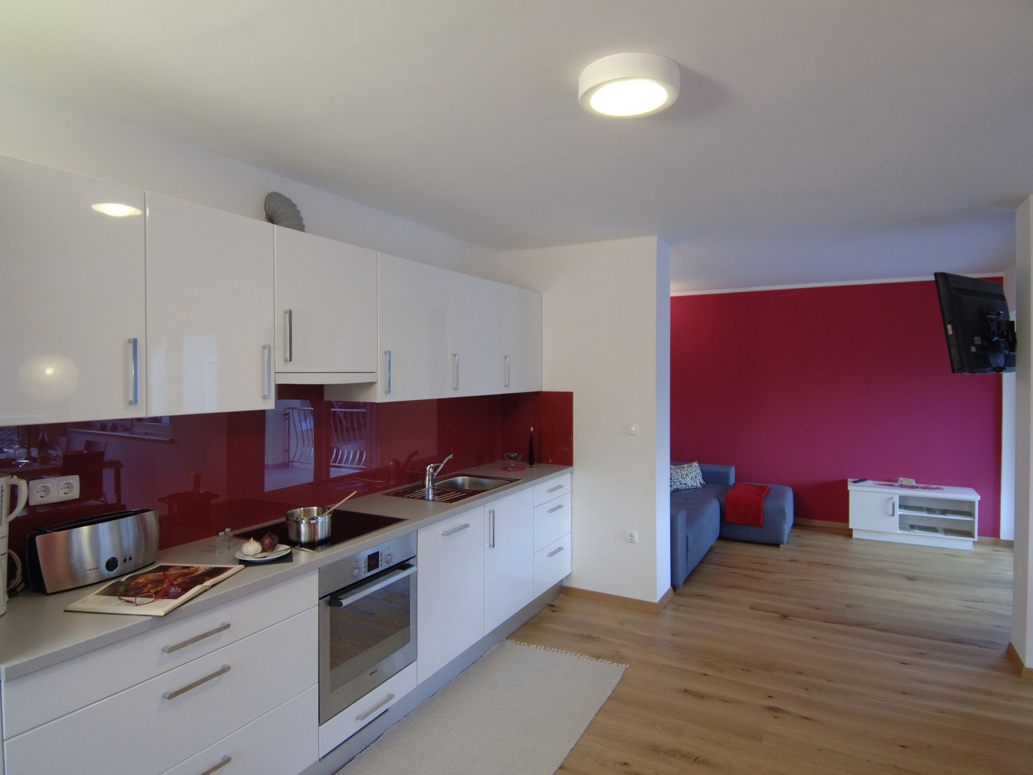 A modern kitchen with white cabinets, a stainless steel oven, and a red backsplash. There is a dining area with a blue sofa and a flat-screen TV mounted on the wall.