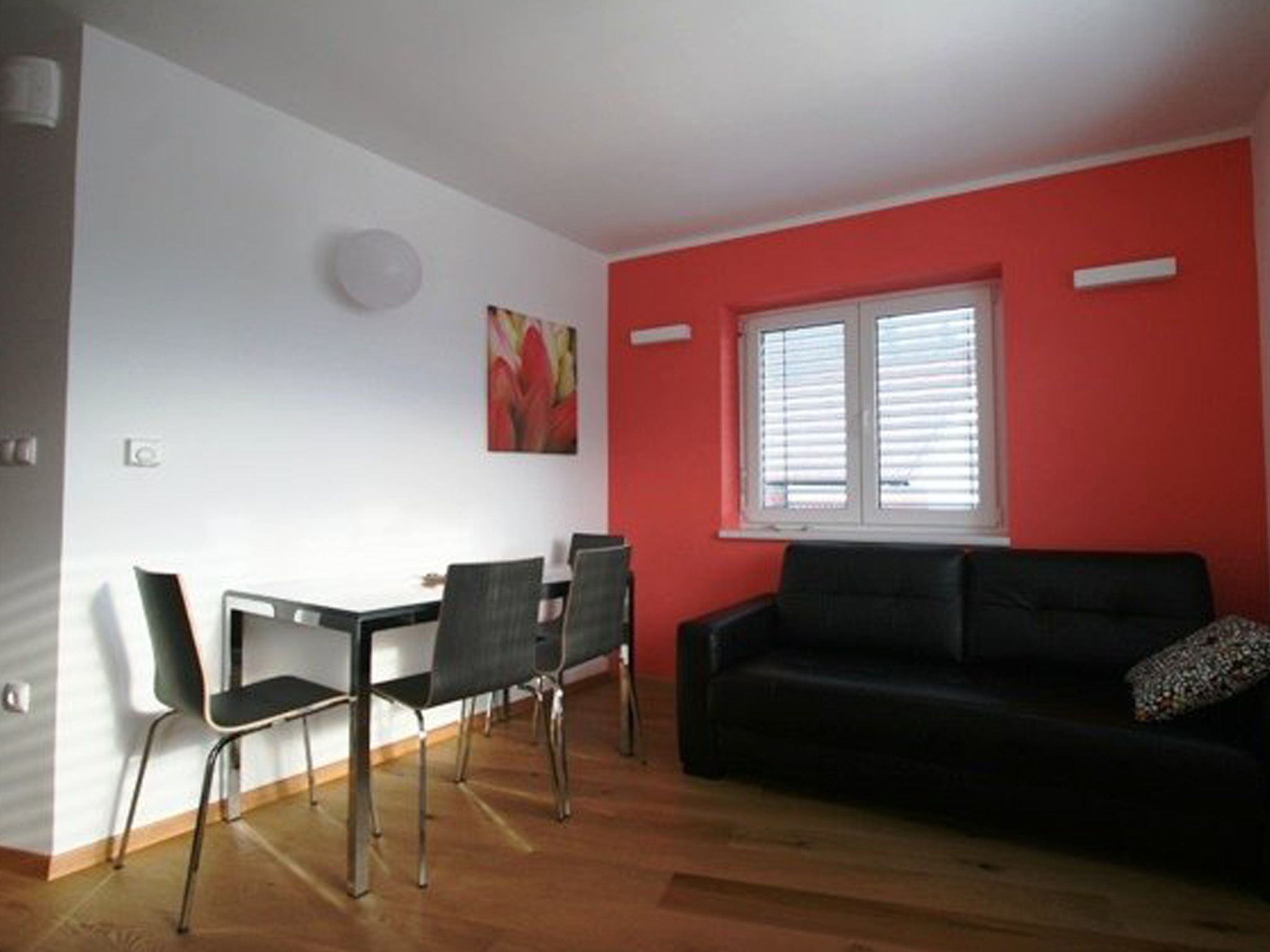 A room with a dining table, four chairs, a black sofa, a window with blinds, and a red accent wall.
