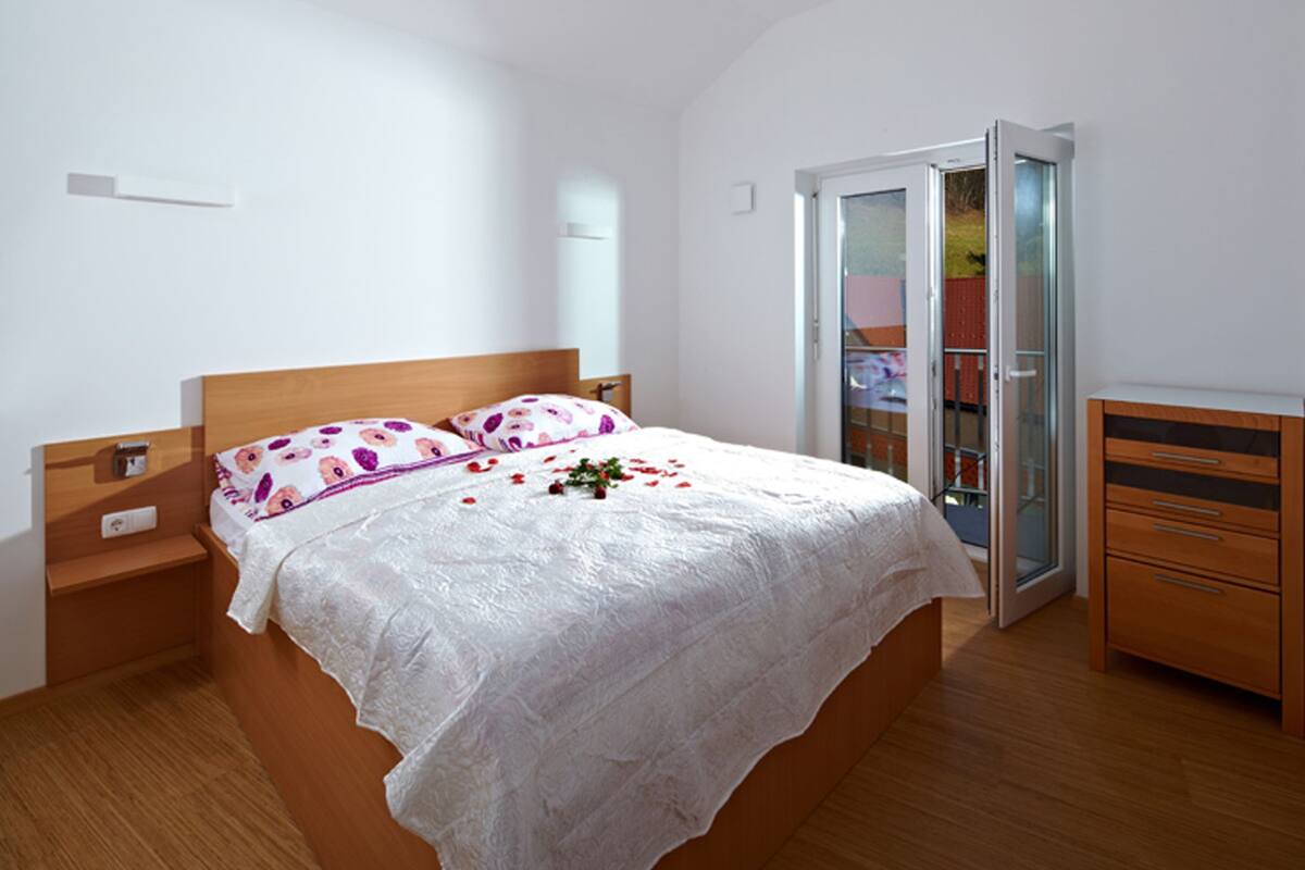 Een slaapkamer met een houten bed, witte lakens, een nachtkastje, een dressoir en een schuifpui die leidt naar een balkon.