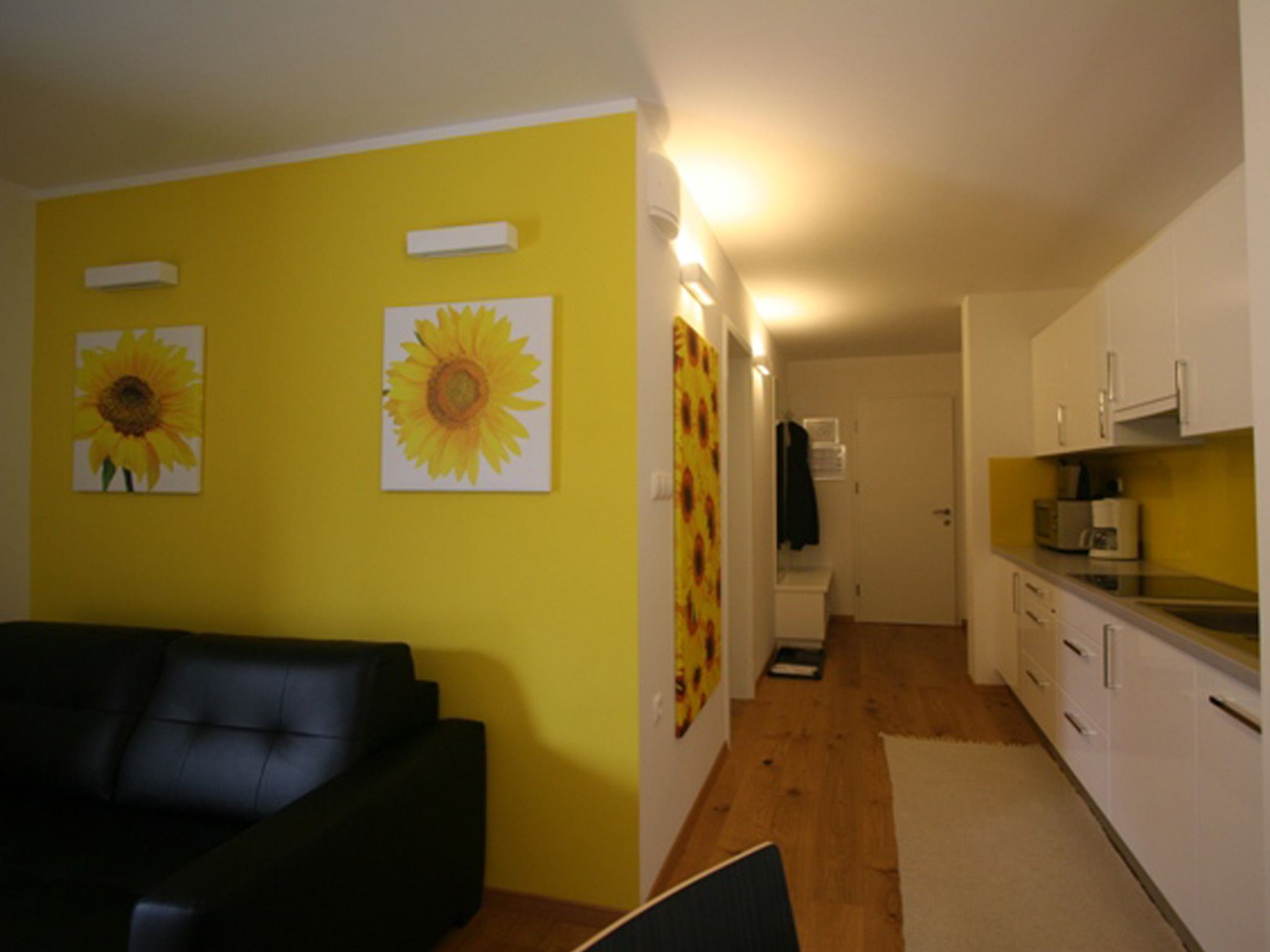 A room with a black sofa, a kitchen with white cabinets, and sunflower artwork on the walls.