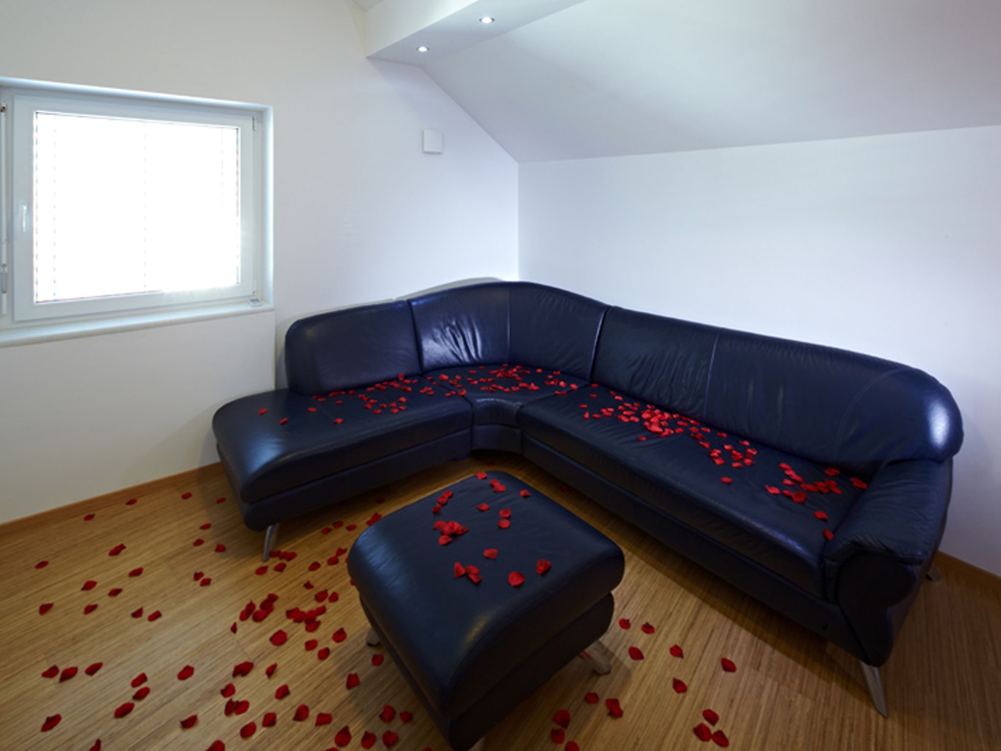 Una habitación con un sofá seccional de cuero negro y un reposapiés, piso de madera y una ventana con persianas.
