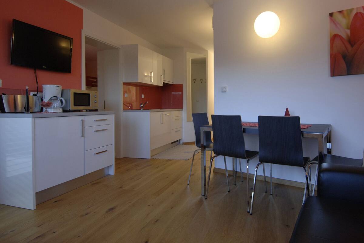 Een moderne keuken met witte kasten, een flatscreen televisie en een eethoek met twee stoelen en een tafel.