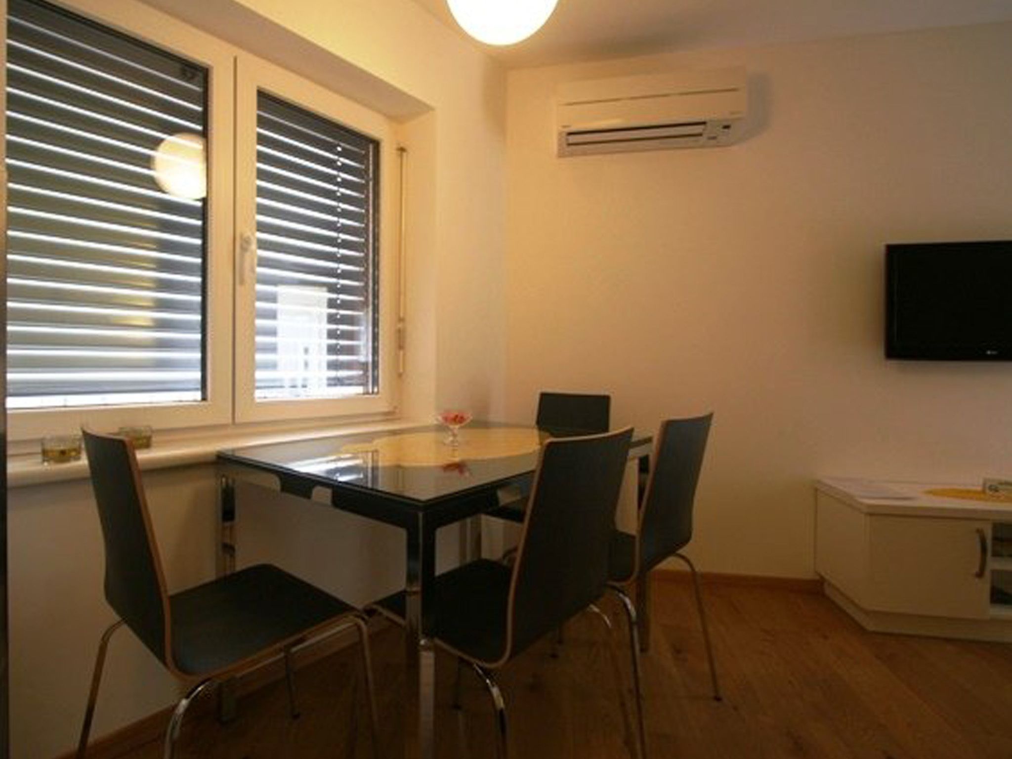 A small dining area with a table and four chairs, a window with blinds, an air conditioning unit, and a television on the wall.