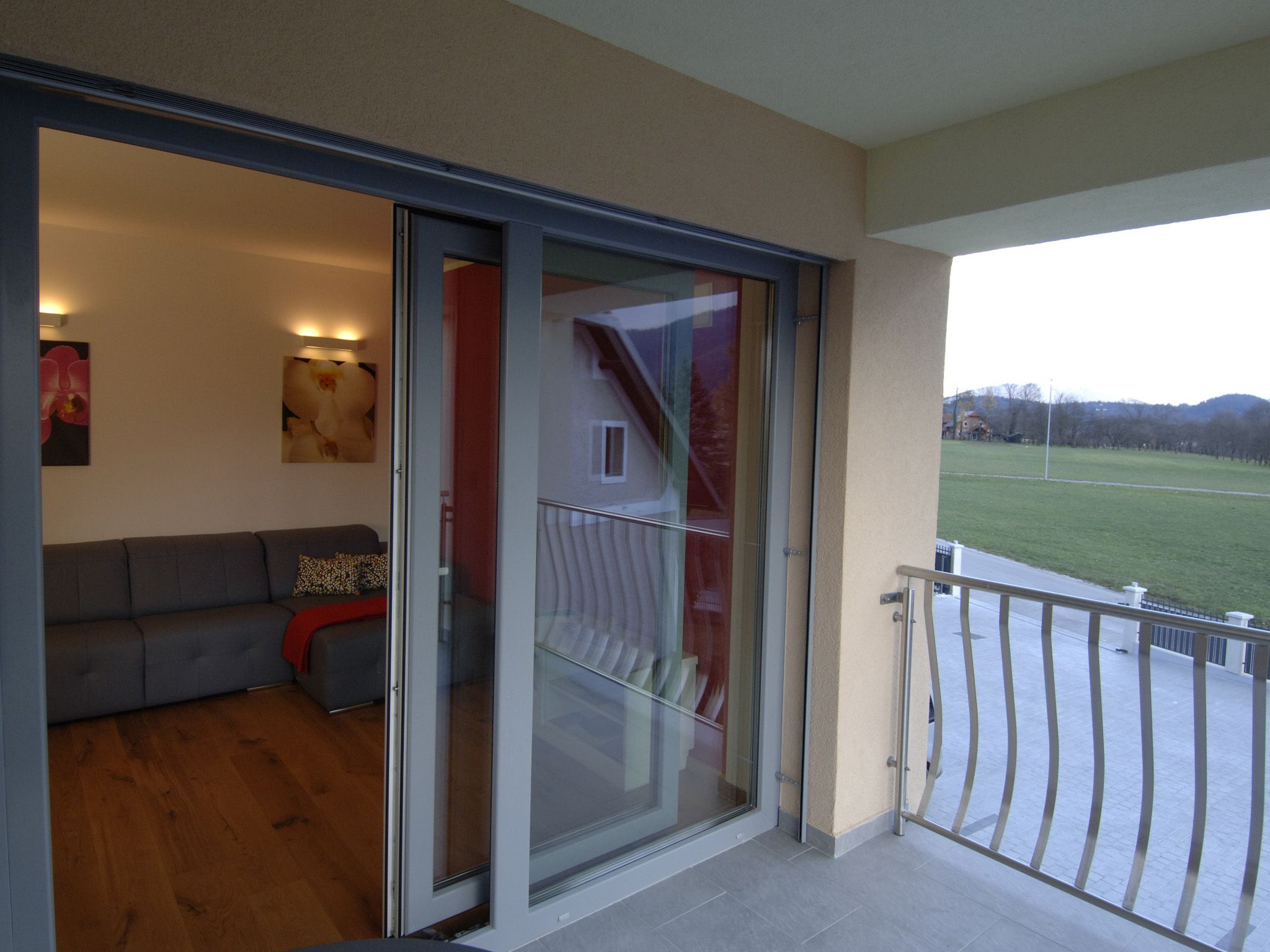 A modern living room with a grey sofa, a red throw pillow, and a glass door leading to a balcony with a metal railing.
