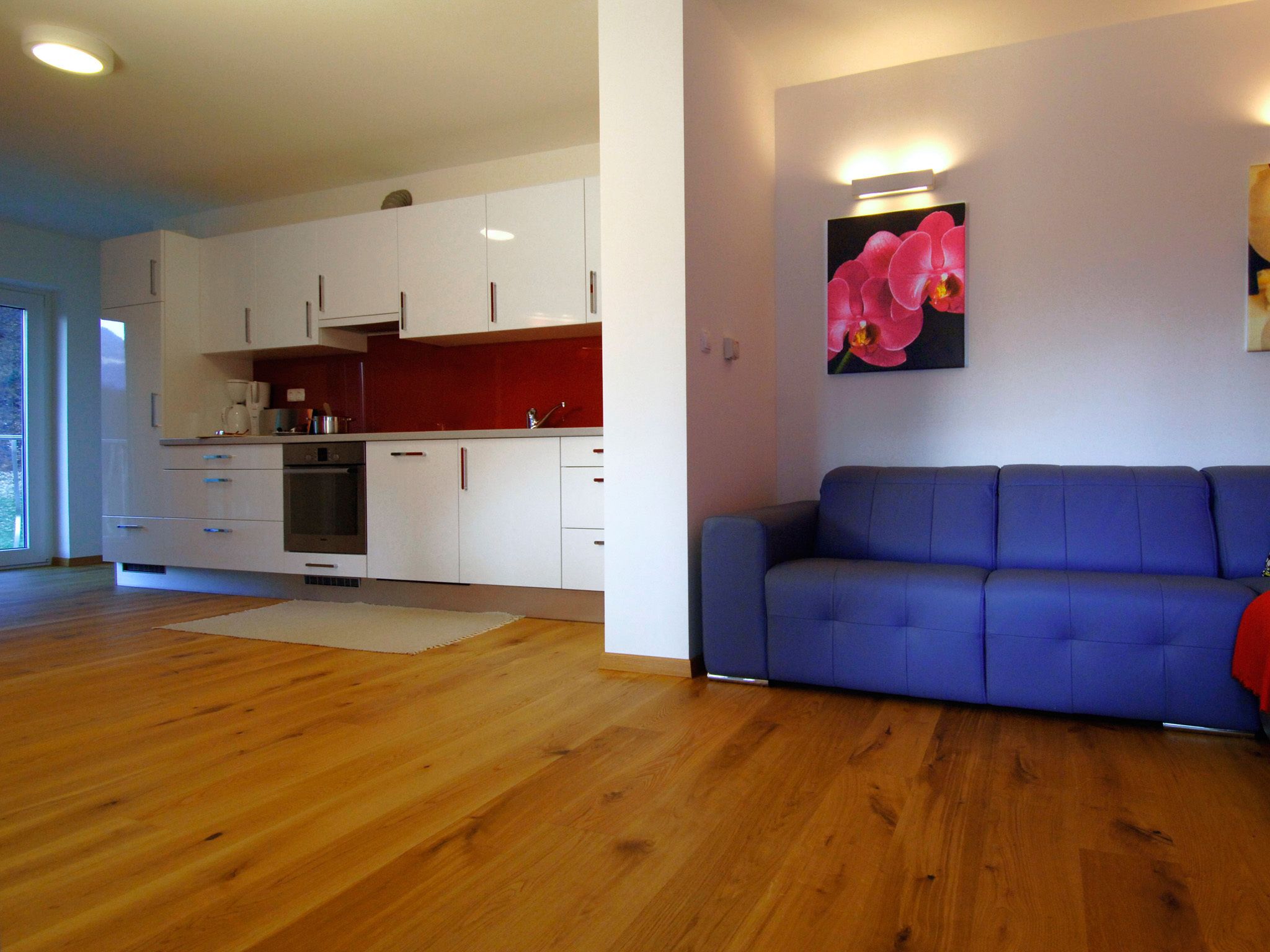 A modern kitchen with white cabinets and a red backsplash, a blue sofa, and a wooden floor.