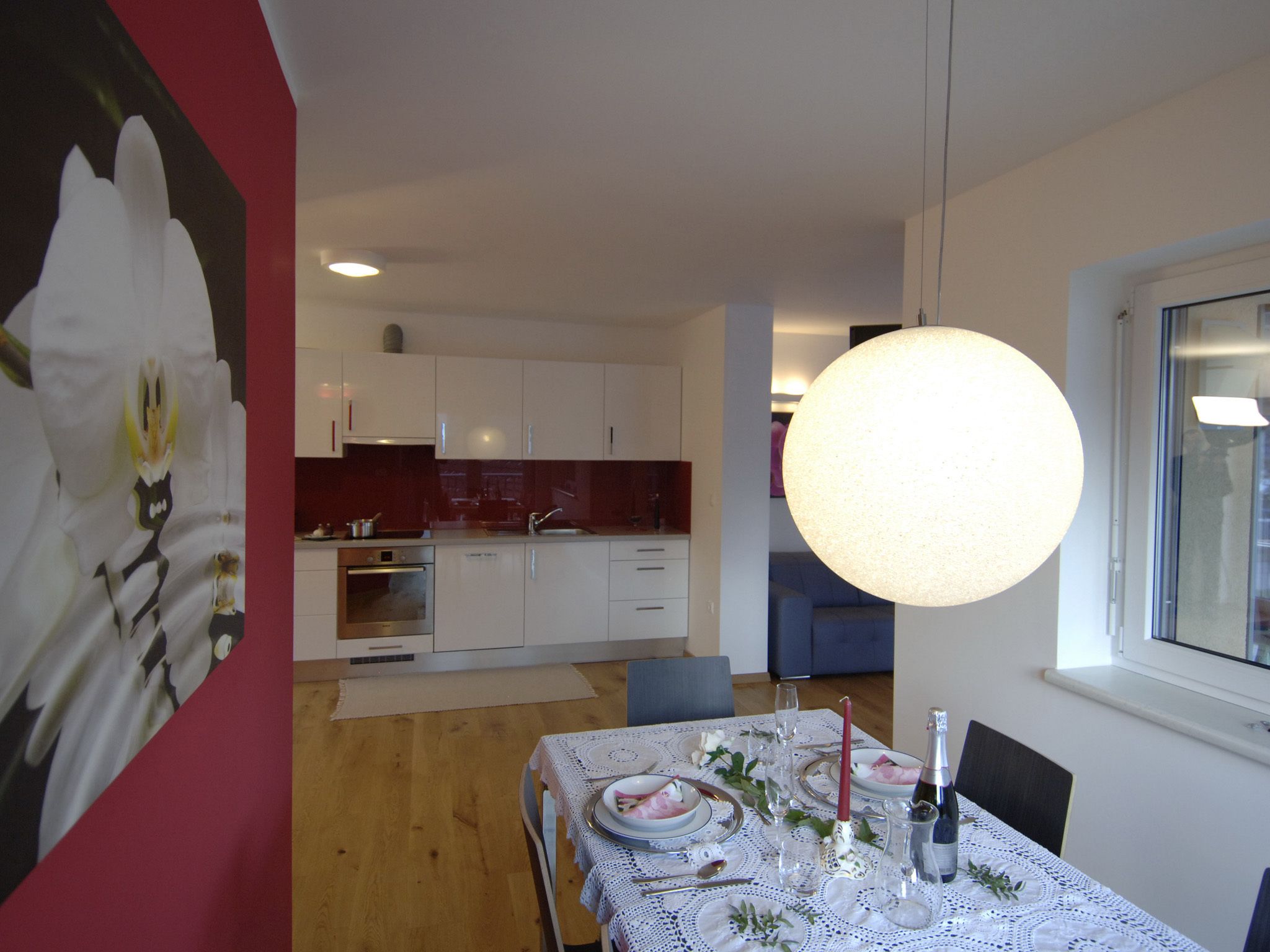 A dining area with a table set for four, a modern kitchen in the background, and a large pendant light above the table.