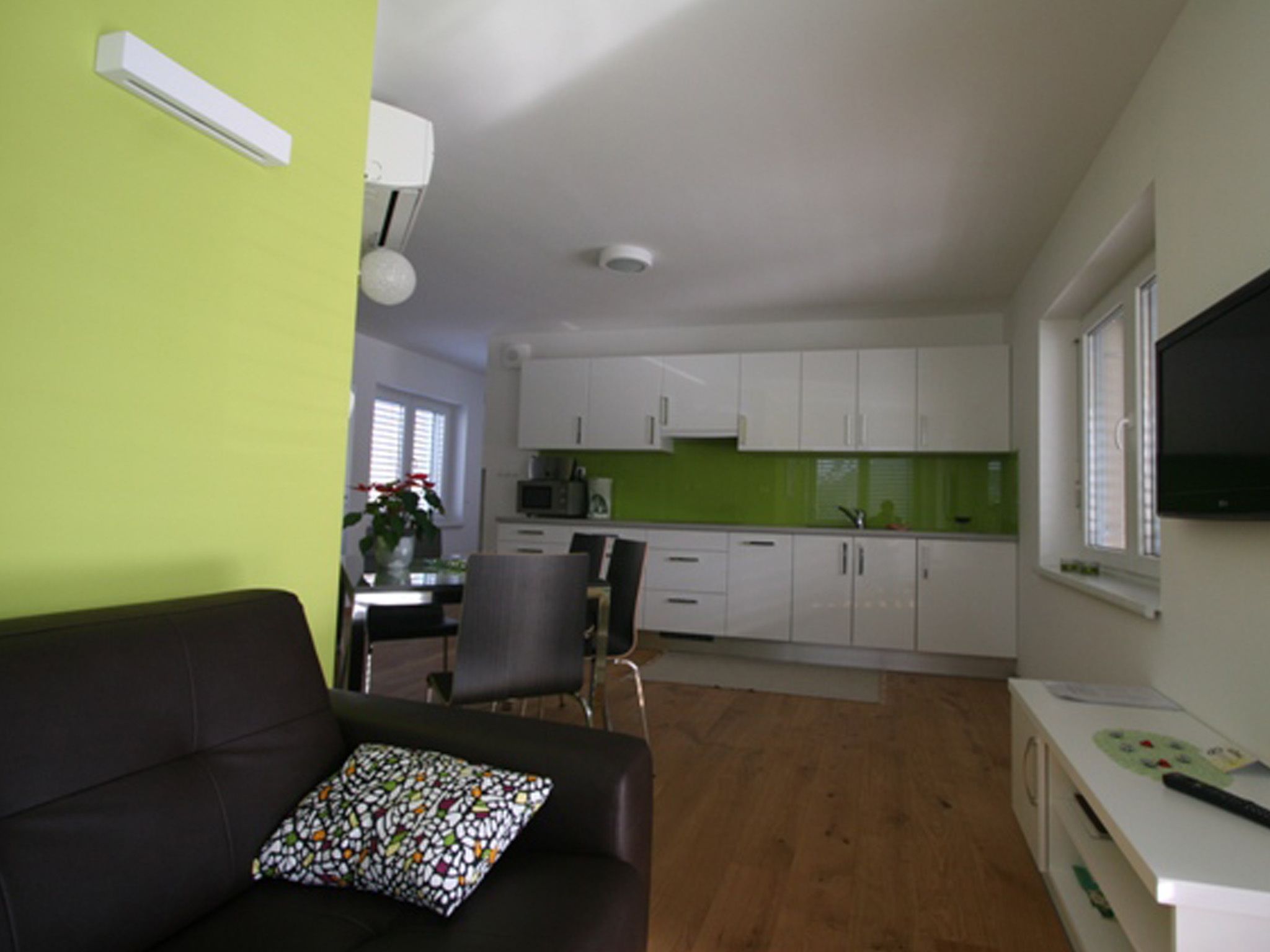 Una sala de estar moderna con un sofá de cuero negro y un cojín colorido, un comedor con mesa y sillas, una cocina con armarios blancos y un salpicadero verde, y una ventana con persianas.