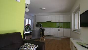 Una sala de estar moderna con un sofá de cuero negro y un cojín colorido, un comedor con mesa y sillas, una cocina con armarios blancos y un salpicadero verde, y una ventana con persianas.