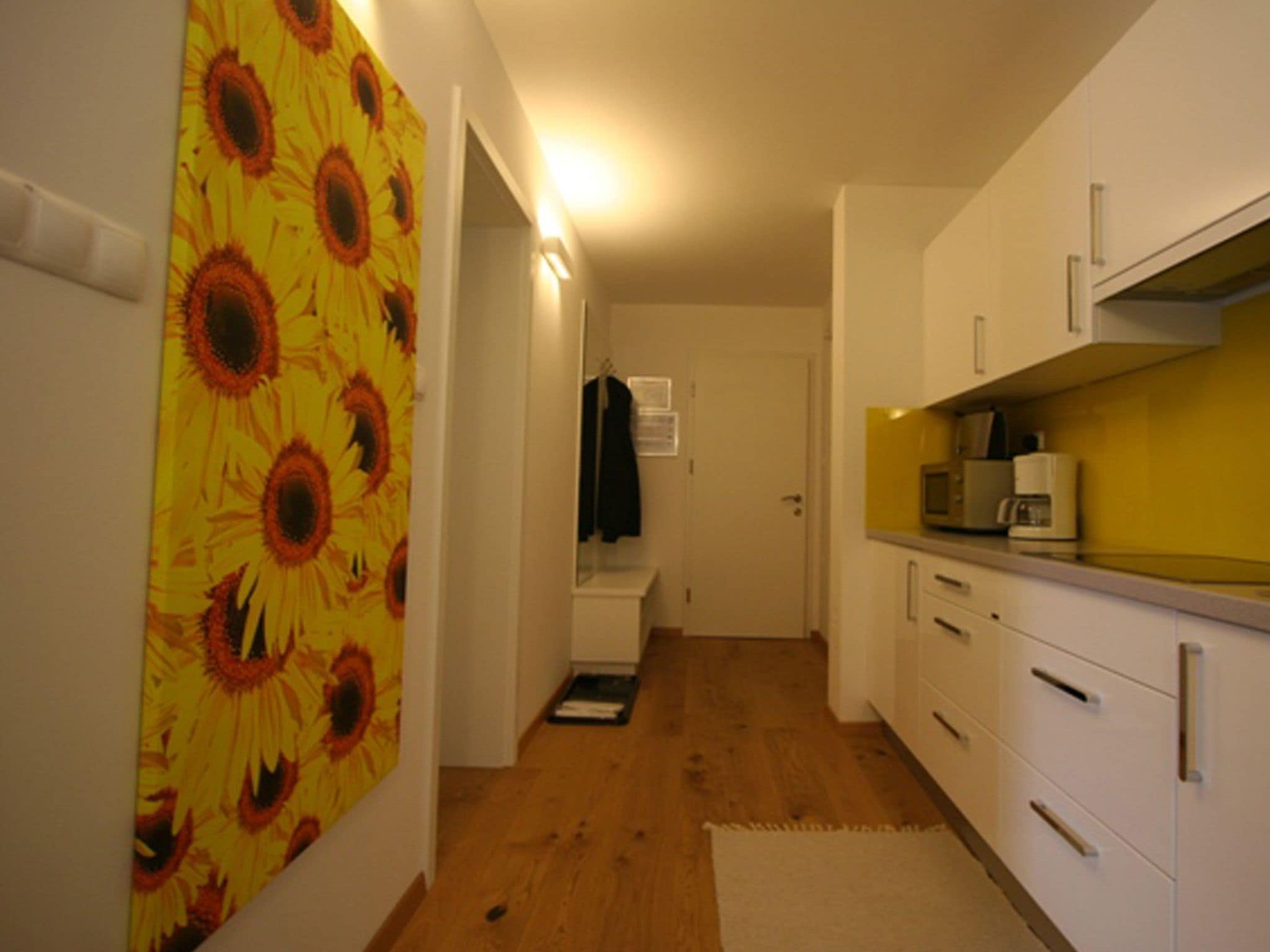 A narrow hallway with wooden flooring, white cabinets, and a yellow wall with sunflower wallpaper.