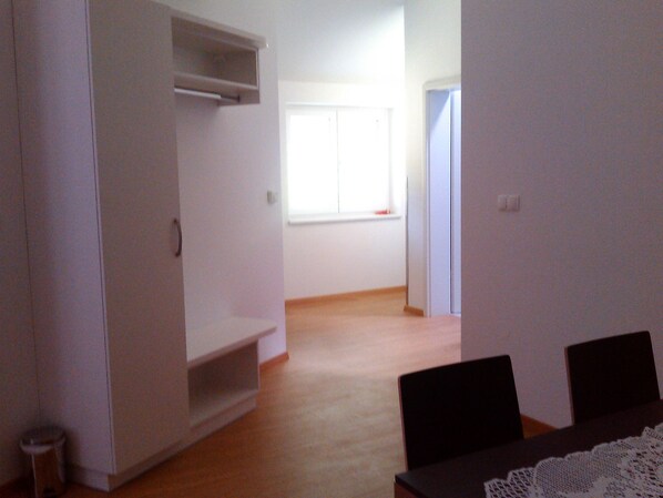 Una habitación con piso de madera, paredes blancas, un ventanal con persianas y una puerta que da a otra habitación.