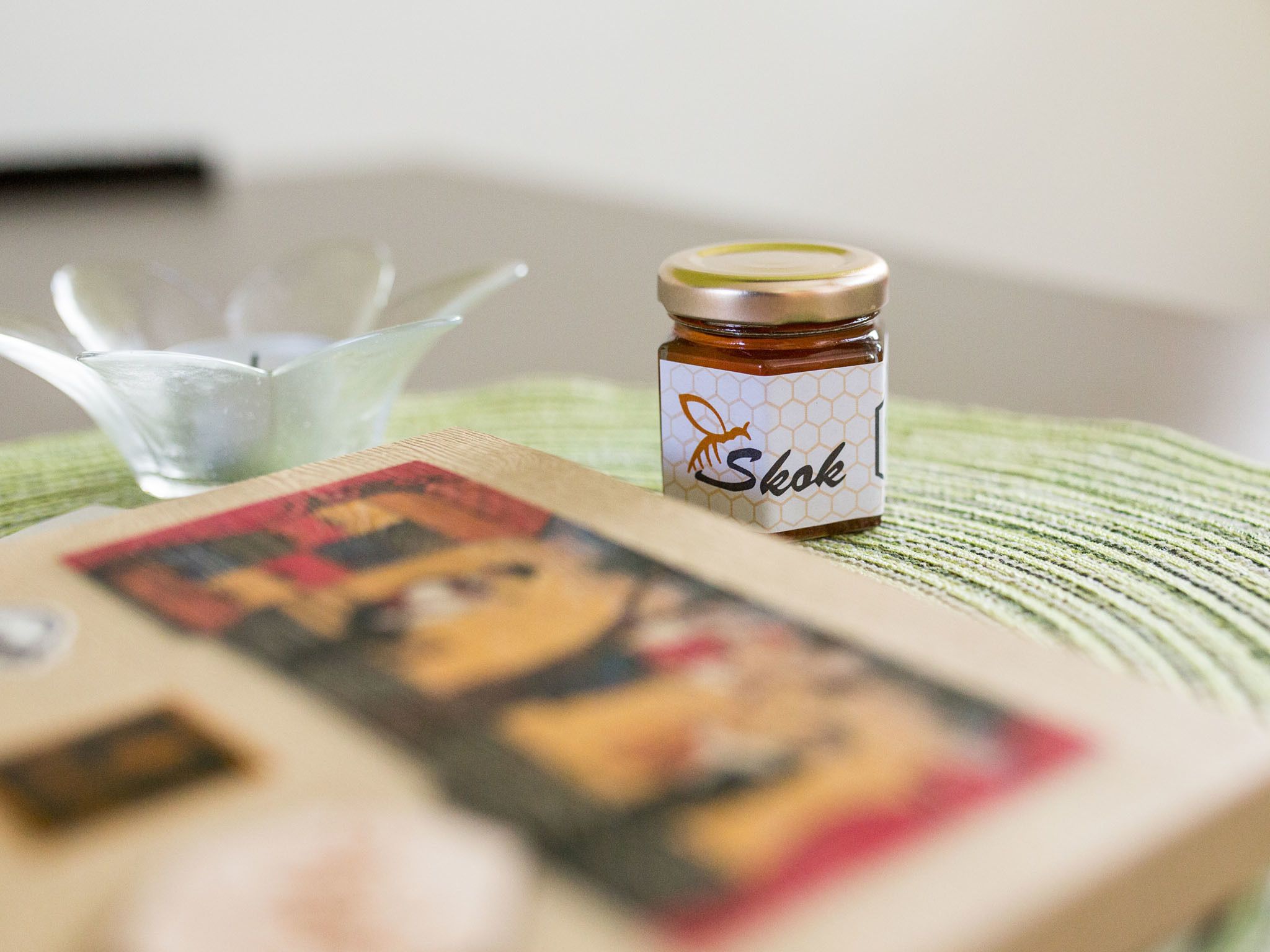 A jar of honey labeled 'Skok' on a table with a clear glass object and a book.