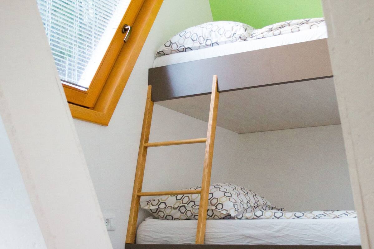 Una cama nido con escalera de madera, ventana con persianas y ropa de cama estampada.