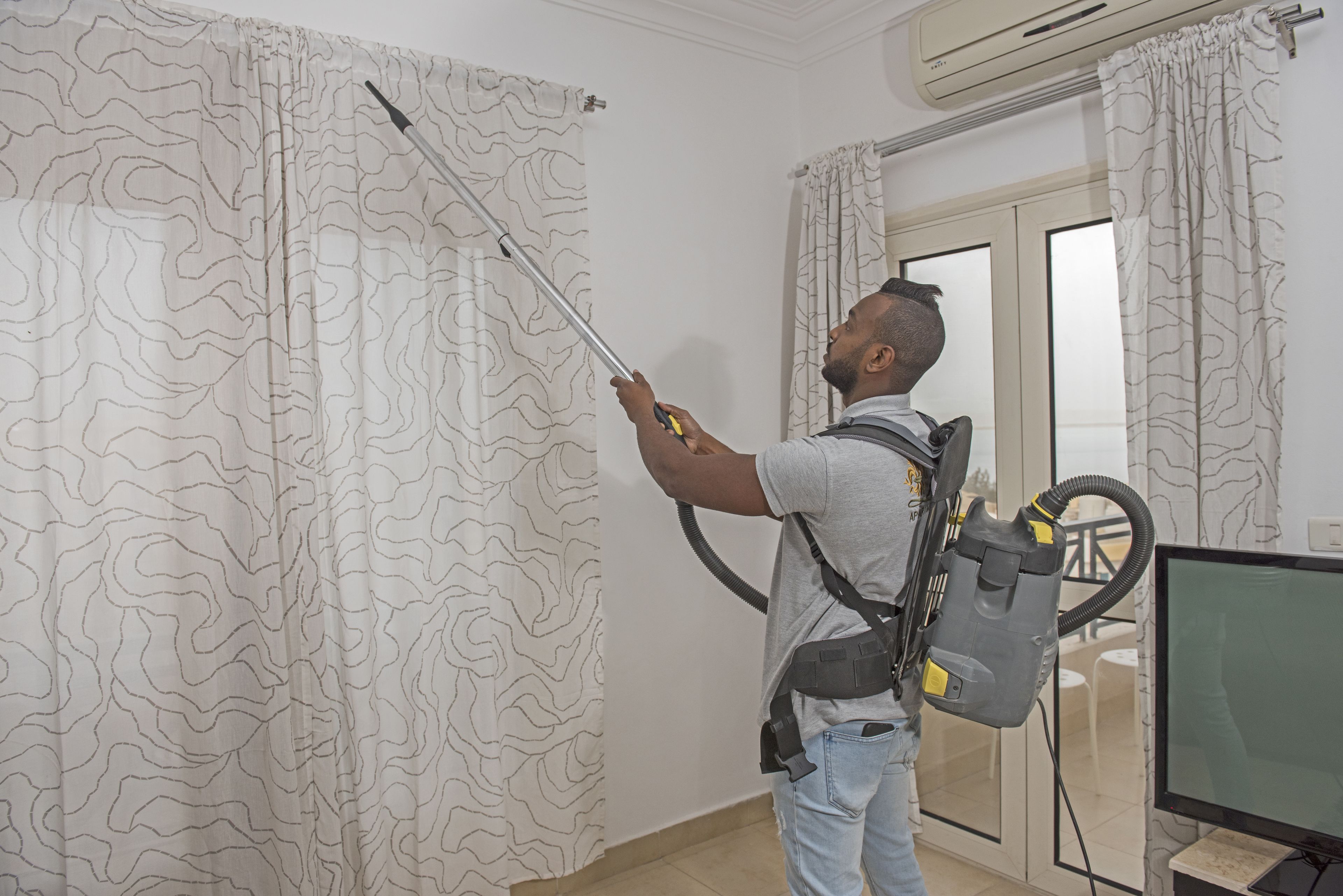 A person using a vacuum cleaner in a room with patterned curtains, a television, and an air conditioning unit.