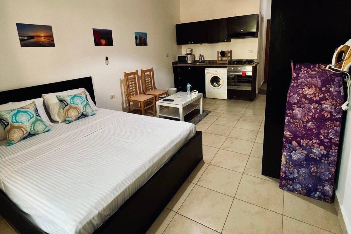 A bedroom with a bed, a desk, a chair, a washing machine, and a kitchen area with cabinets and a microwave.