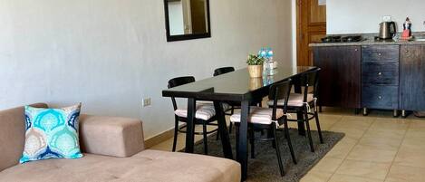 A modern living room with a sofa, a coffee table, a dining table with chairs, a kitchen area with cabinets, and a hanging light fixture.