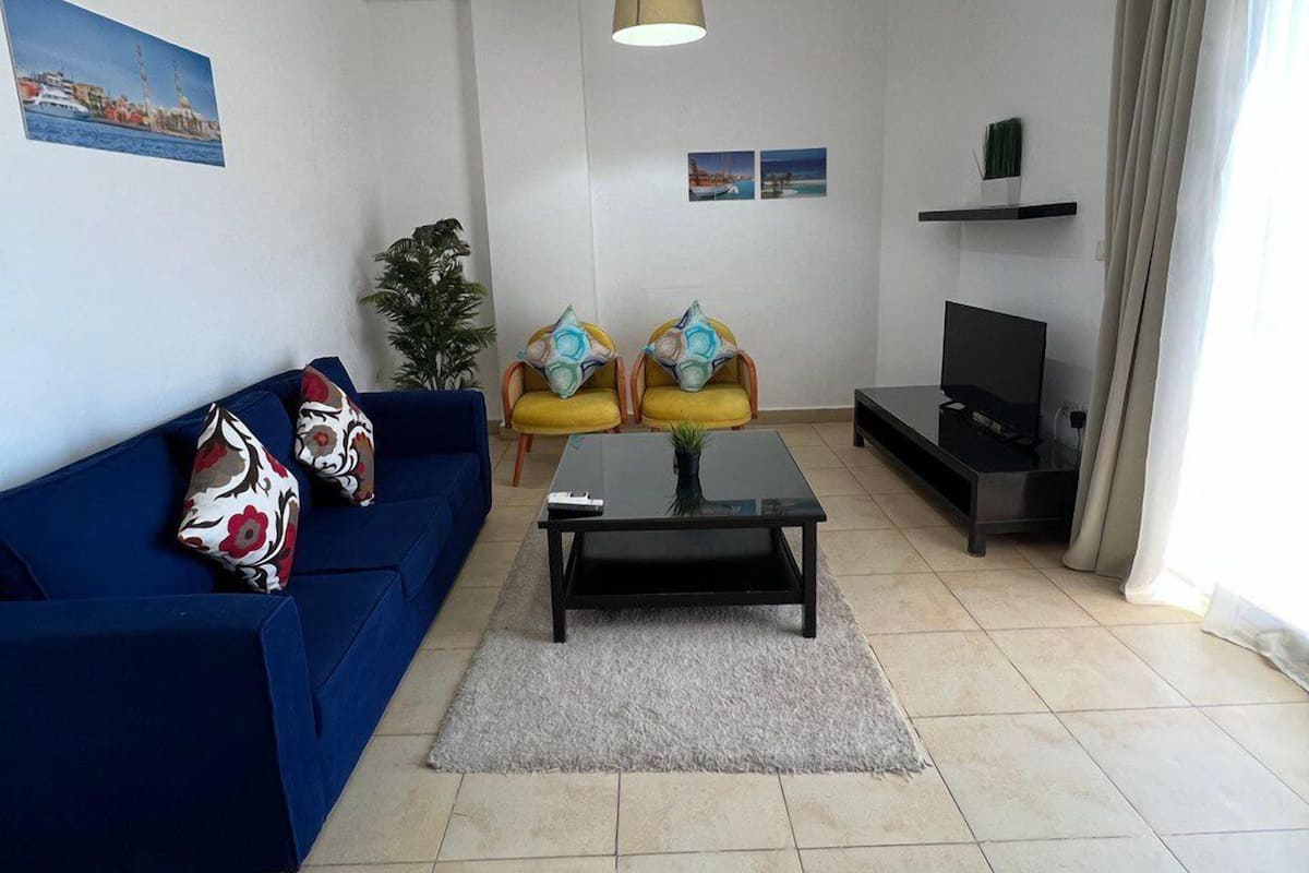 A living room with a blue sofa, two yellow armchairs, a coffee table, a TV stand, and a painting on the wall.