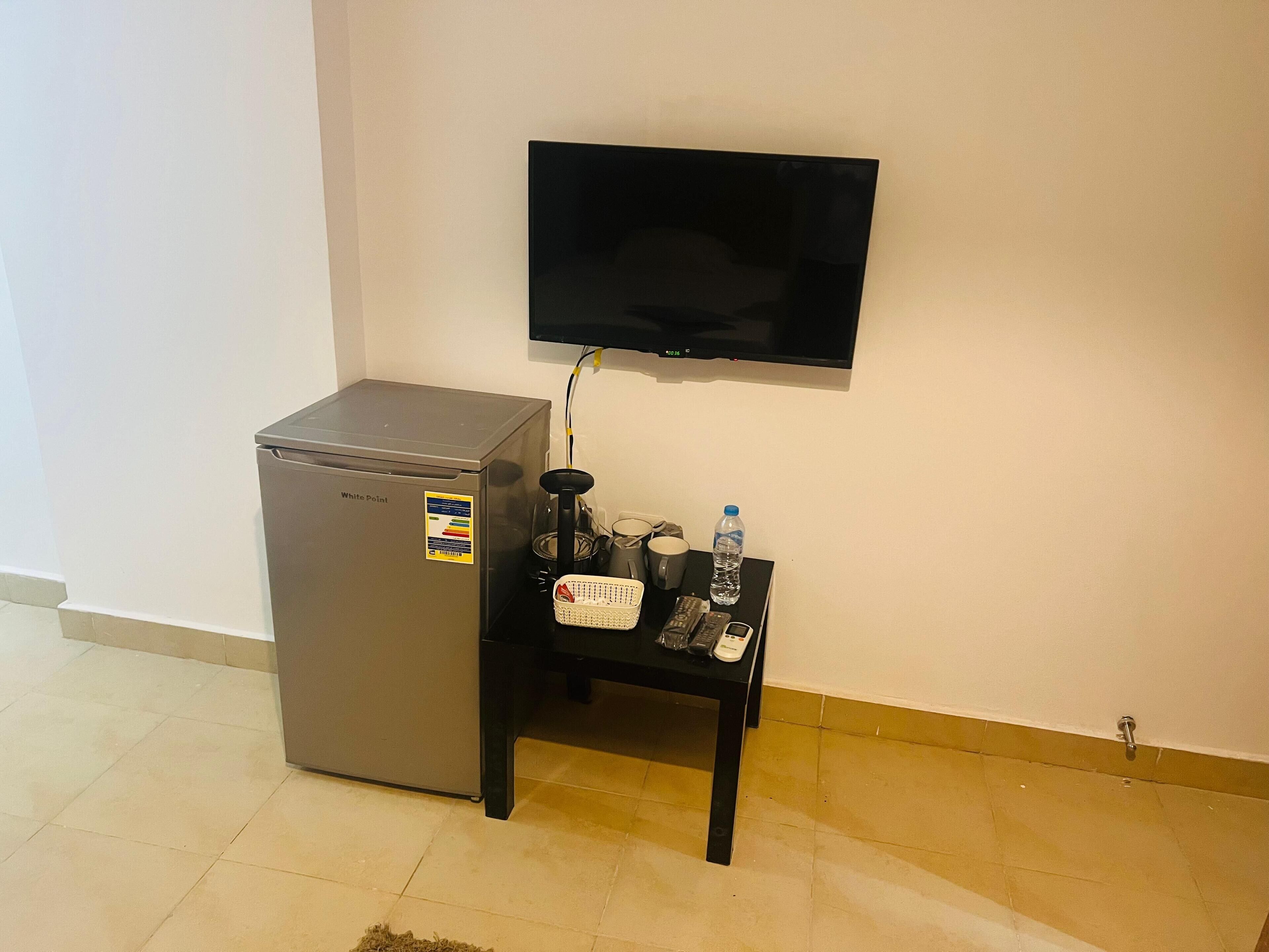 A compact refrigerator, a small table with a water bottle, a remote, and a mobile phone.