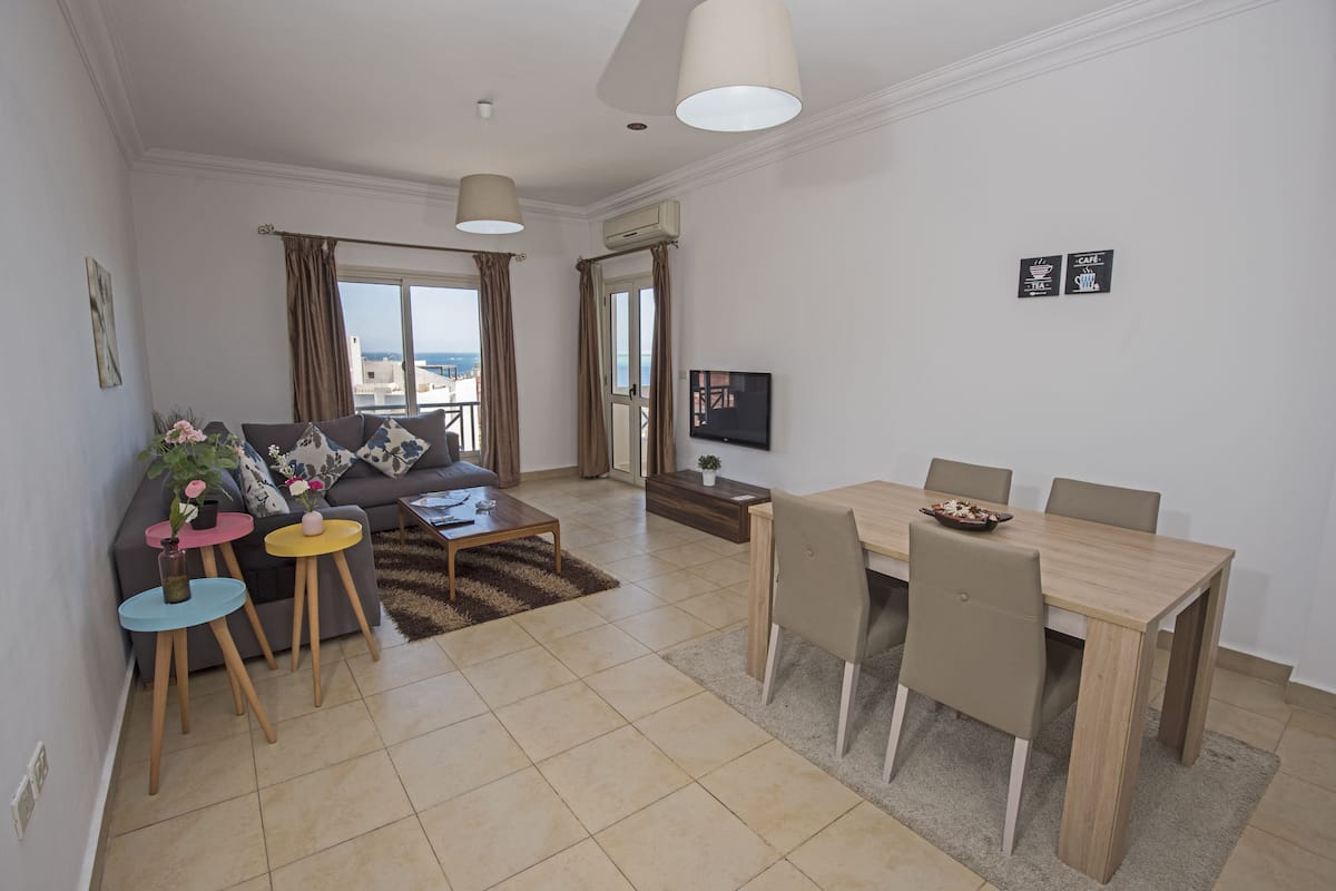 A modern living room with a sofa, coffee table, dining table, and chairs. There is a TV on a stand, a window with curtains, and a view of the sea.