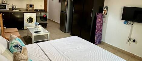 Une chambre d’hôtel comprenant un lit, une petite table avec une chaise, un lave-linge, un réfrigérateur, une télévision et un coin cuisine avec des placards et des appareils électroménagers.