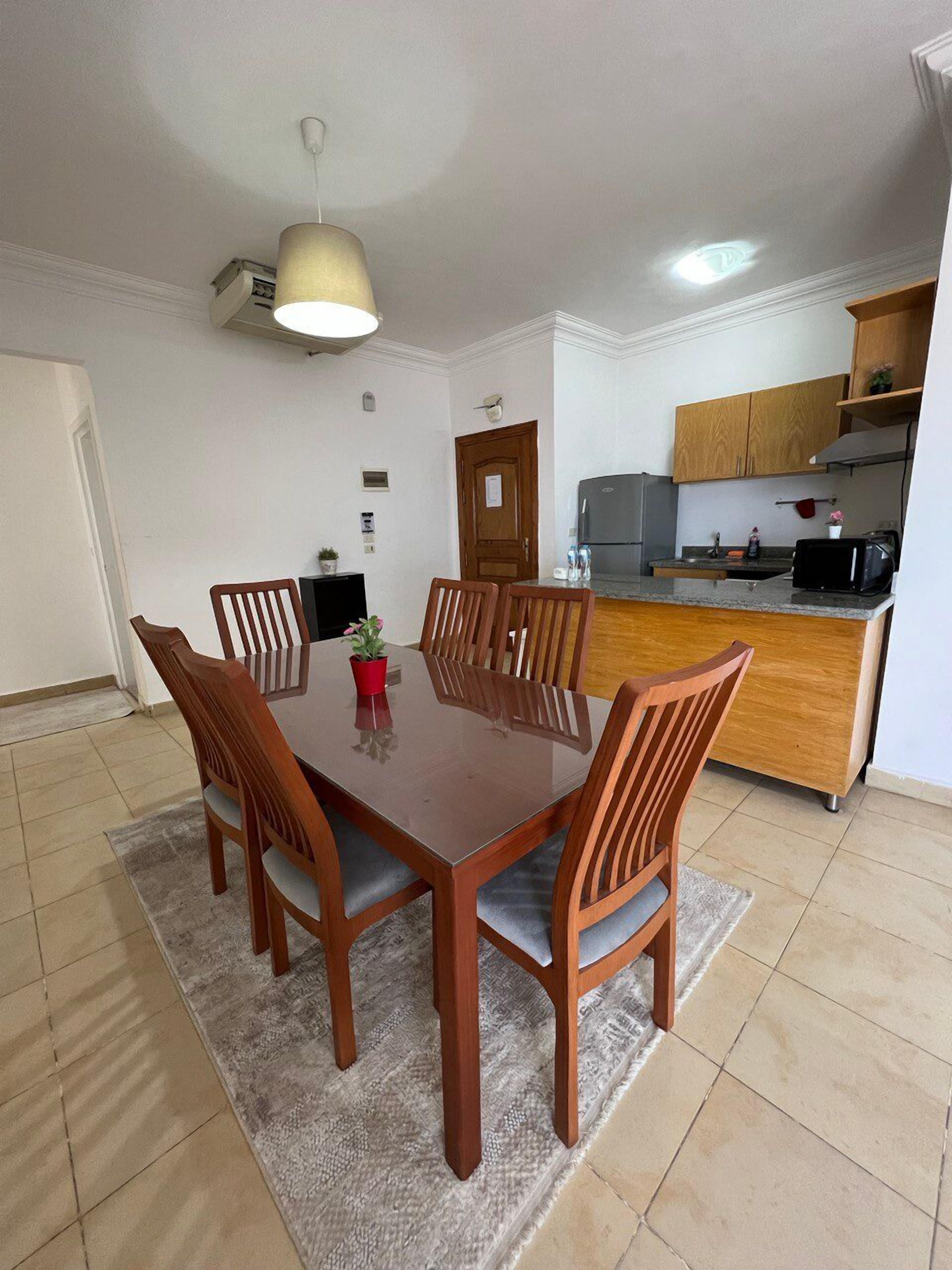 A dining area with a wooden table and chairs, a small potted plant, and a kitchen in the background.