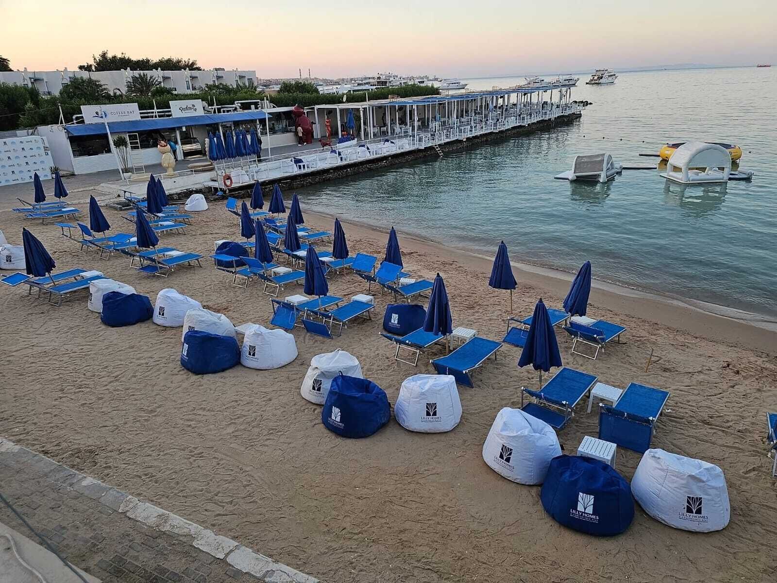 Une plage bordée de rangées de chaises longues et de parasols bleus, une jetée avec des bateaux, et un bâtiment sur lequel on peut lire l’inscription « Coffee Bay ».