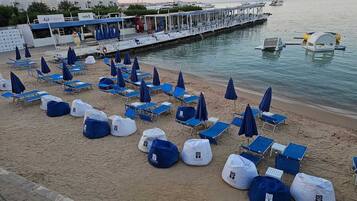 Une plage bordée de rangées de chaises longues et de parasols bleus, une jetée avec des bateaux, et un bâtiment sur lequel on peut lire l’inscription « Coffee Bay ».