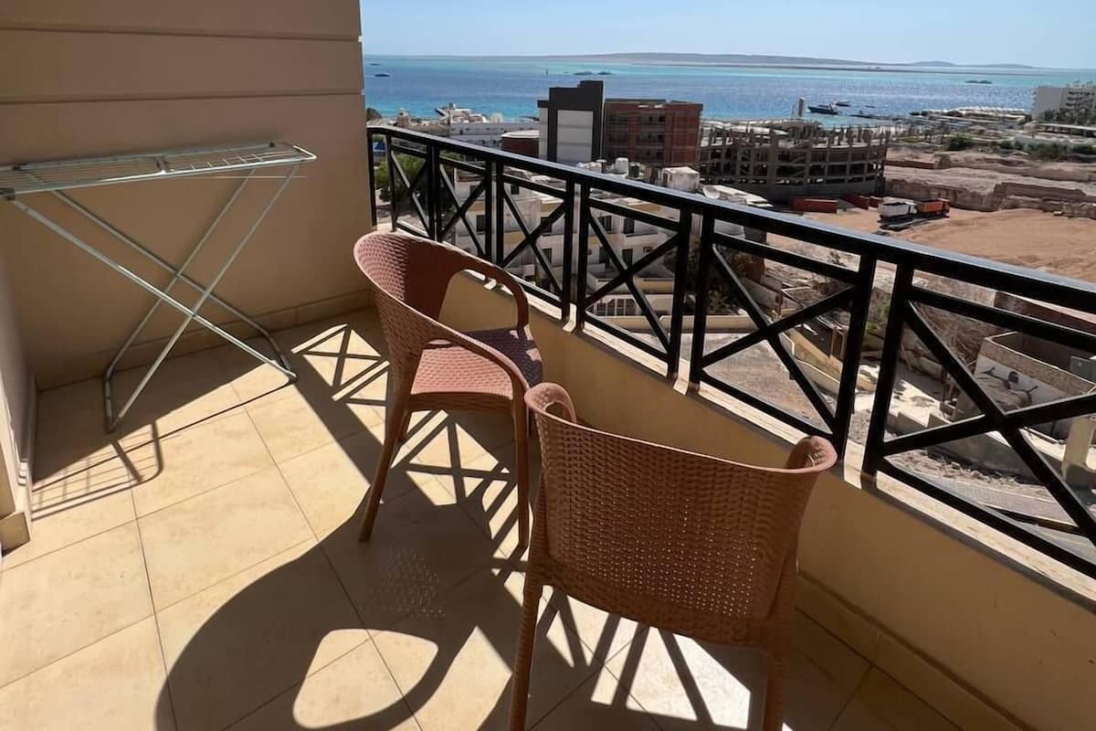 A balcony with two wicker chairs and a metal drying rack. The balcony overlooks a coastal area with buildings and boats.