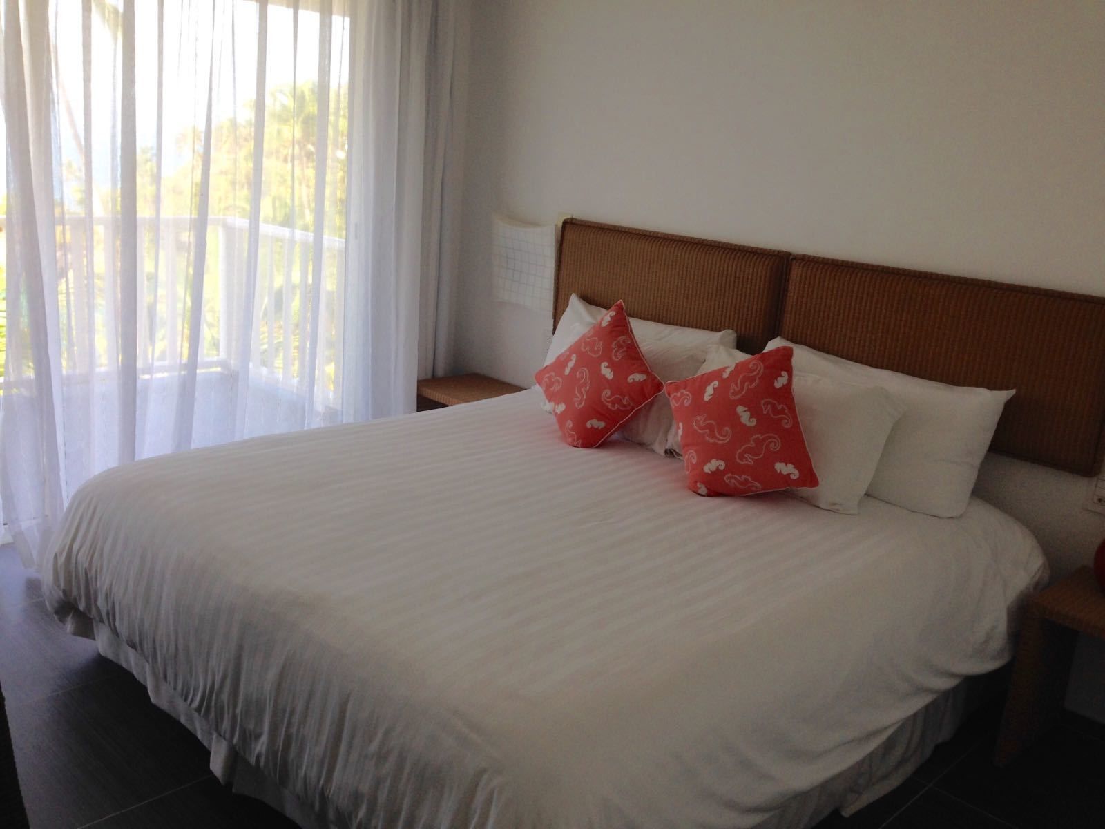 Cama bem arrumada com roupa de cama branca e almofadas vermelhas, cabeceira de madeira e janela com cortinas leves.