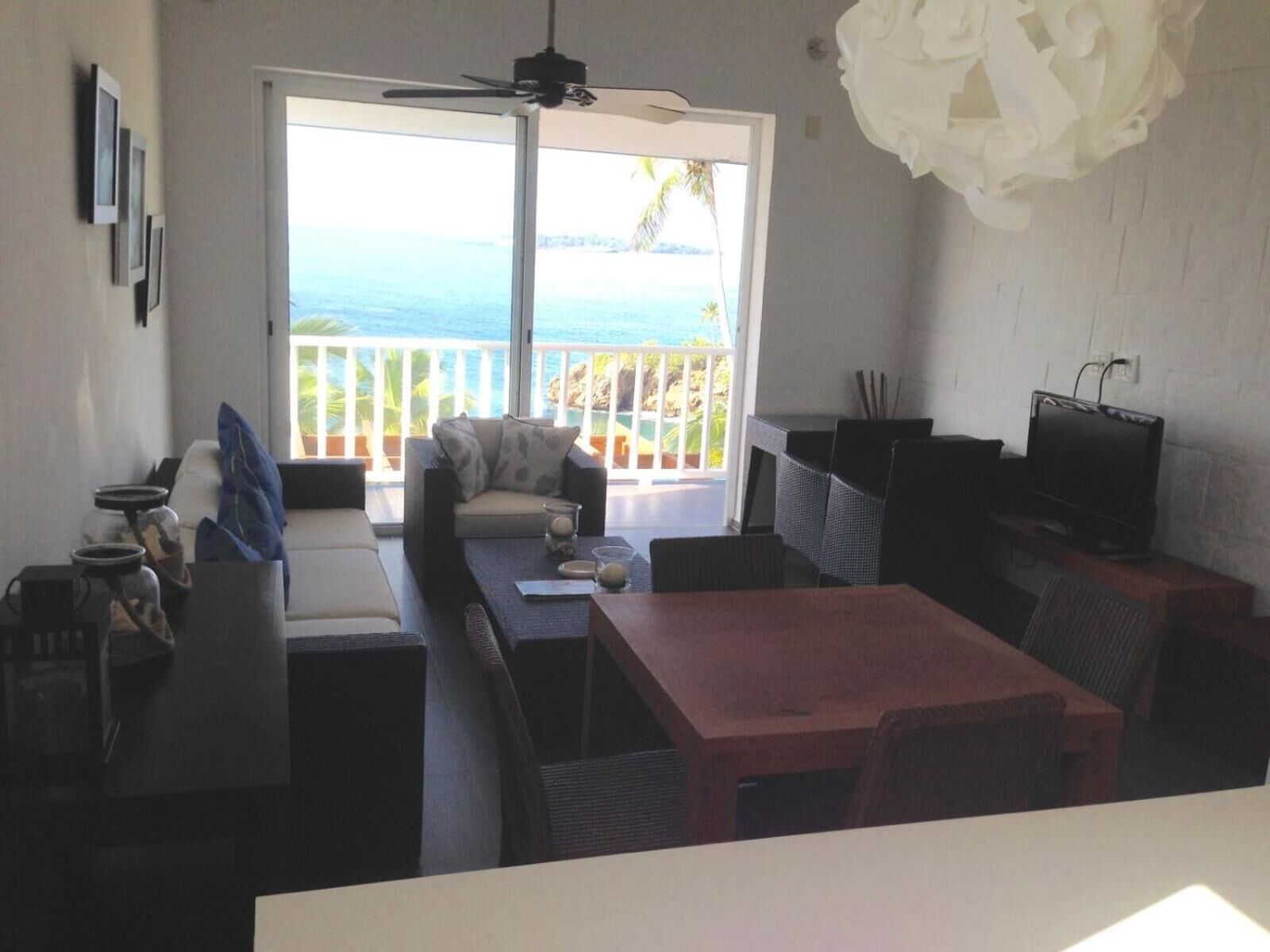A living room with a sofa, a table, and a TV. There is a balcony with a view of the ocean.
