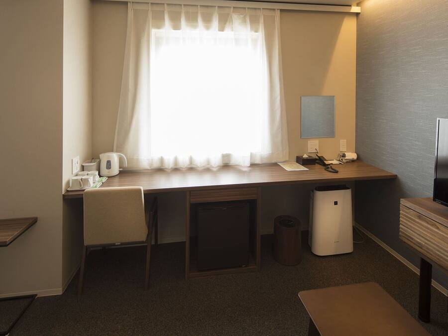 Habitación de hotel con escritorio, silla, cubo de basura pequeño, cafetera, televisor y ventana con cortinas.
